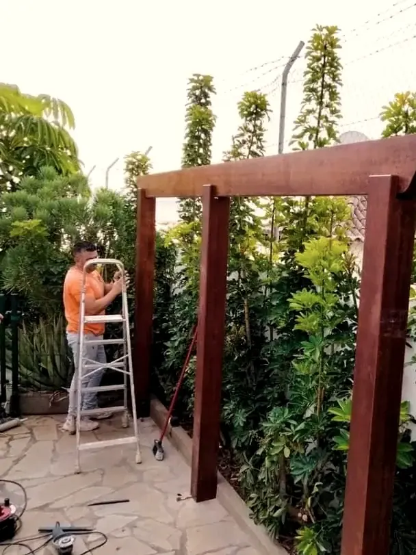 Operario montando una pérgola de madera en una zona ajardinada con setos